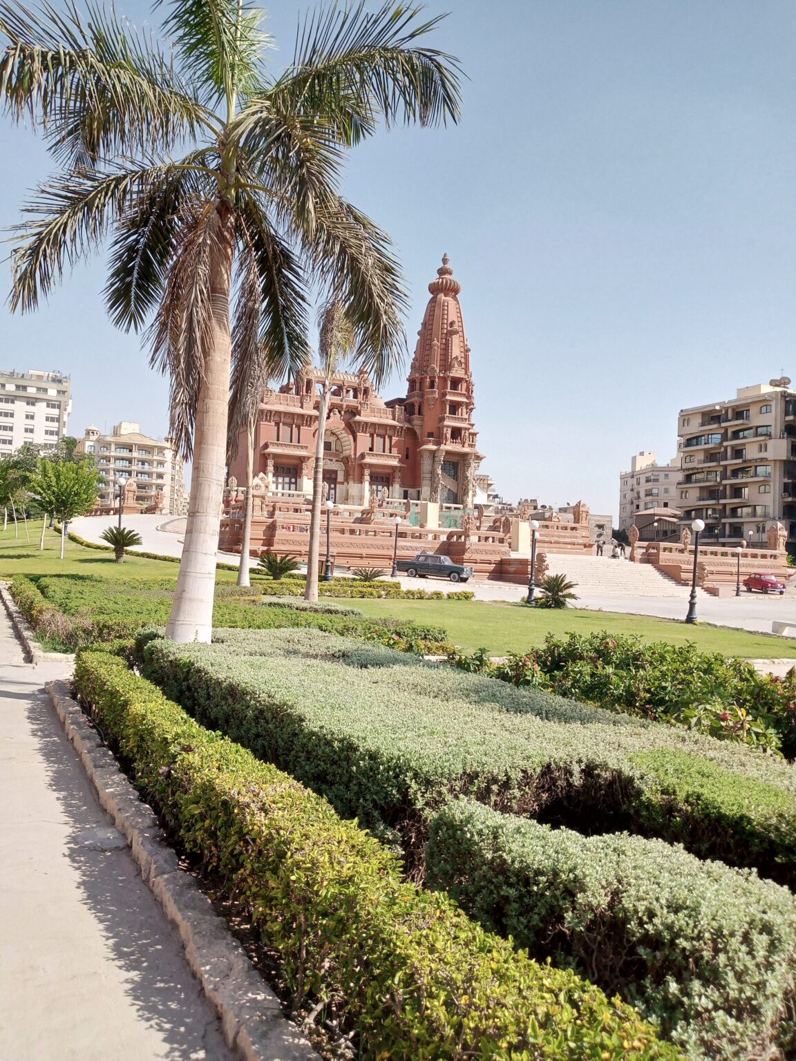 صورة لقصر البارون إمبان في حي مصر الجديدة بالقاهرة، يظهر القصر بطرازه المعماري الفريد المستوحى من المعابد الهندوسية والبوذية، محاط بحدائق خضراء وممرات حجرية، وتغمره إضاءة نهارية تبرز تفاصيله الزخرفية.