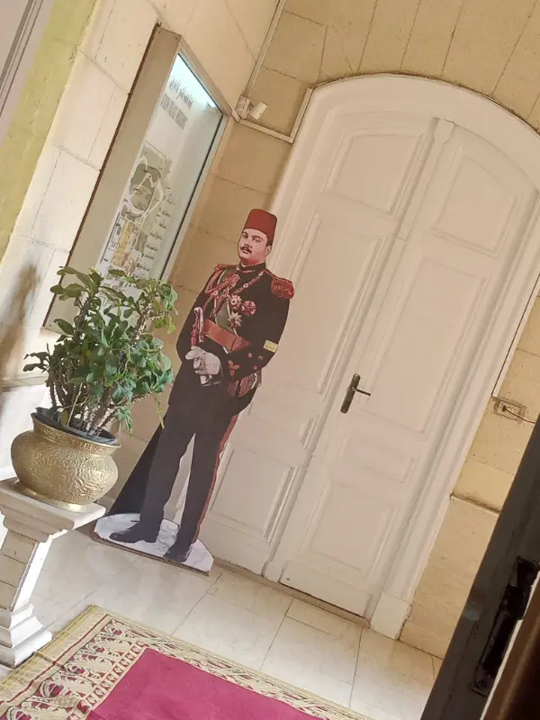 "مجسم بالحجم الطبيعي للملك فاروق داخل قصر عابدين، يرتدي زيًا عسكريًا أسود مزخرف بالذهب مع طربوش أحمر وسيف، في مدخل مزين بالسجاد الأحمر والديكور الكلاسيكي."