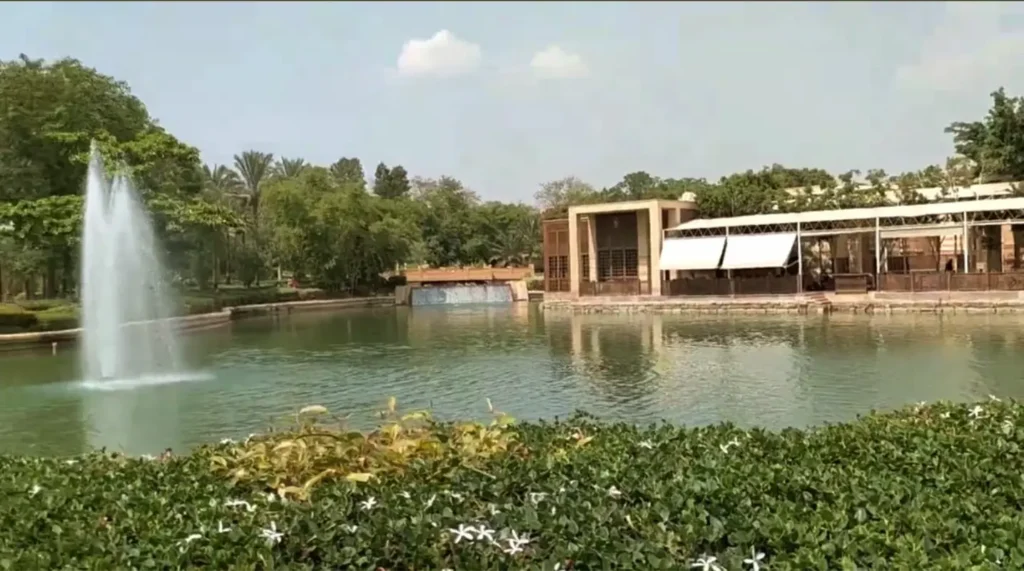 منظر لحديقة الأزهر في القاهرة يظهر نافورة مياه وسط بحيرة صغيرة محاطة بالأشجار والزهور، مع مبنى حديث في الخلفية، يعكس أجواء هادئة ومكان مناسب للتنزه والاسترخاء.