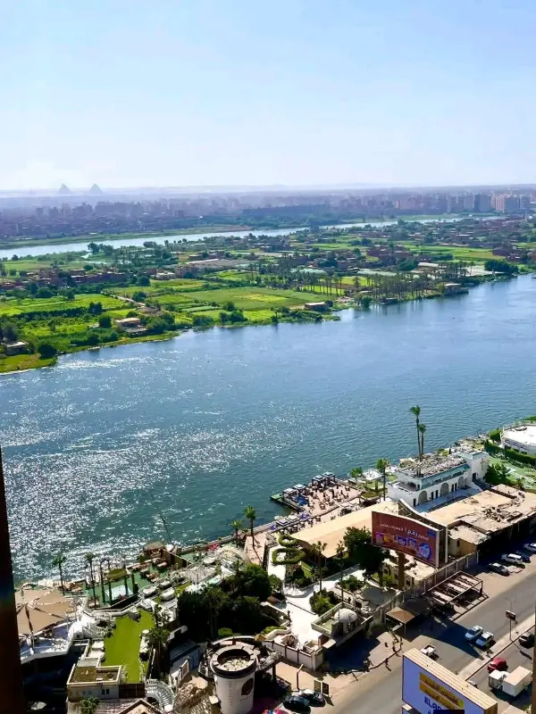 مشهد لكورنيش النيل في القاهرة يظهر النهر الواسع مع مراكب ودكّاكين على ضفافه، تحيط به مساحات خضراء ومبانٍ حضرية، وفي الخلفية أفق المدينة مع الأهرامات البعيدة.