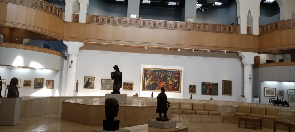 قاعة داخل متحف الفن المصري الحديث تعرض منحوتات على قواعد دائرية ولوحات فنية متنوعة على الجدران، مع شرفة خشبية مزخرفة وإضاءة تبرز جمال الأعمال الفنية.