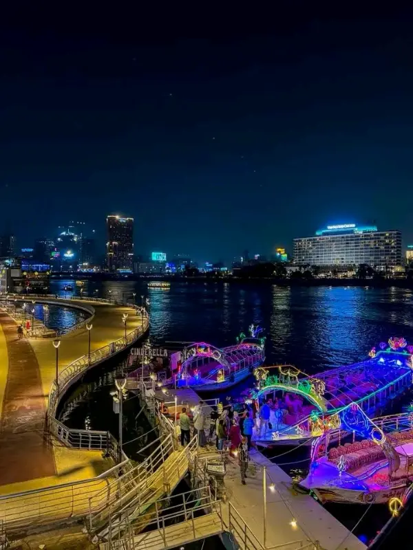 ممشى أهل مصر ليلاً مع قوارب مضاءة بألوان زاهية، أضواء المدينة تنعكس على مياه النيل، وأجواء احتفالية على الواجهة النيلية.